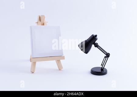 Wooden Easel Miniature with Blank White Square Canvas for Artists and Painters - Mockup. Mini Wooden Stand with Clean Artboard and Small Black Table Lamp on White Background, Copy Space Stock Photo