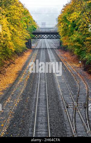 Narrow city bridge across multiline Railway road along the hills with yellow autumn trees with fallen leaves shrouded in a viscous autumn mist leading Stock Photo