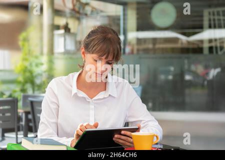 A businesswoman using her digital tablet in a cafe with a cup of coffee and some books and folders on the table Stock Photo