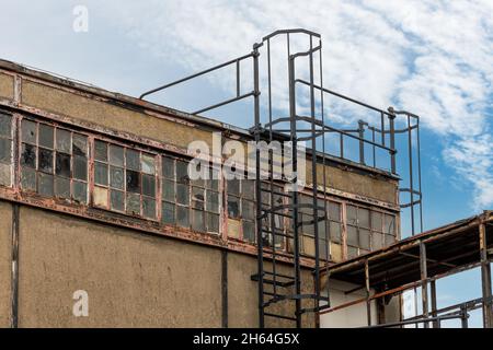 Partial view of facade of dilapidated factory building with iron escape ladder with cage leading to the roof; dirty wall and  windows Stock Photo