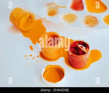 On a white table there is a white palette stained with paint, and next to it there are jars of gouache in red and yellow, one of which is spilled. Cre Stock Photo