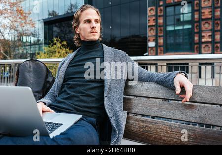 Handsome young man typing on laptop while sitting outdoors on a bench. Concept of work life balance. Businessman freelancer working remotely. Hipster Stock Photo