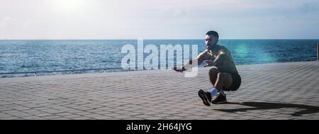 Fit young man doing pistol squats on seafront. Fitness training in morning outdoors. Workout during lockdown outside the gym. Stock Photo