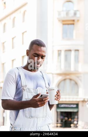 afro man walking the streets and using his smart phone. He is dressing casual summer clothes and holds an reusable cup of coffee Stock Photo