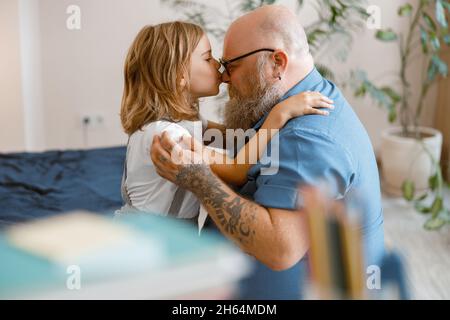 Short haired little girl hugs and kisses happy father nose sitting on bed at home Stock Photo