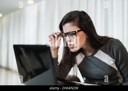 Latin woman with vision problems trying to read online content on a laptop at home. Stock Photo