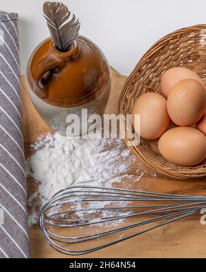 Composition with whisk, flour, eggs and a bird feather in a ceramic container Stock Photo