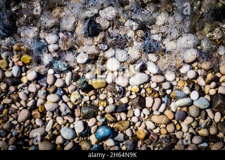 Background of multicolored sea pebbles on the beach in sea foam. Small color wet stones gravel texture background. Stock Photo