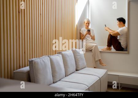 Couple using smartphones on windowsill at home Stock Photo