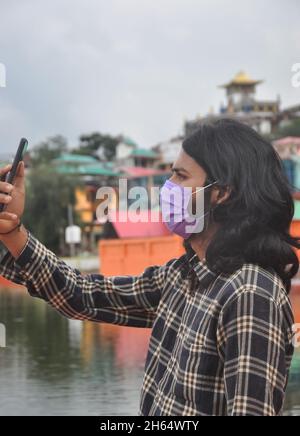 Side view of a long haired young man taking selfie or having video call with his mobile phone with wearing face mask while standing outside Stock Photo