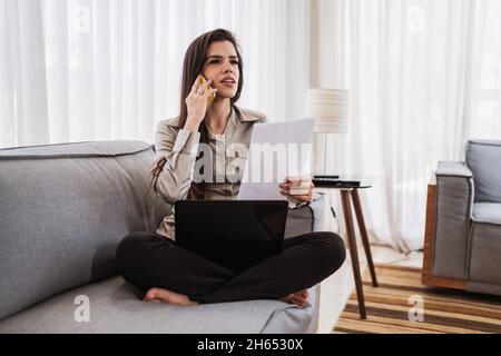 Young beautiful Brazilian woman using smartphone and working with laptop while sitting at sofa, working from home concept. Stock Photo