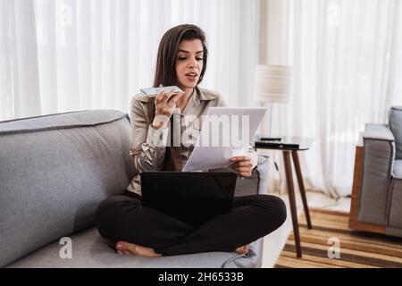 Young beautiful Brazilian woman sending audio message on smartphone and working with laptop while sitting at sofa, working from home concept. Stock Photo