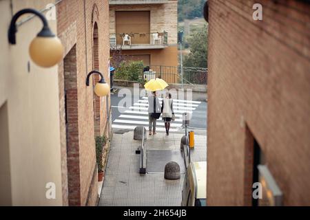 A young couple is crossing the street while walking the city in a gloomy atmosphere on a rainy day. Walk, rain, city, relationship Stock Photo