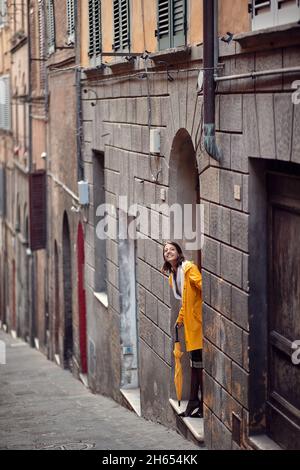 A young girl in a yellow raincoat is standing in the building entrance and checking if it is raining. Walk, rain, city Stock Photo