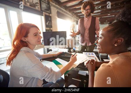 young caucasian beardy guy standing in front of the desk with two female multiethnic colleagues sitting,  looking each other, smiling Stock Photo