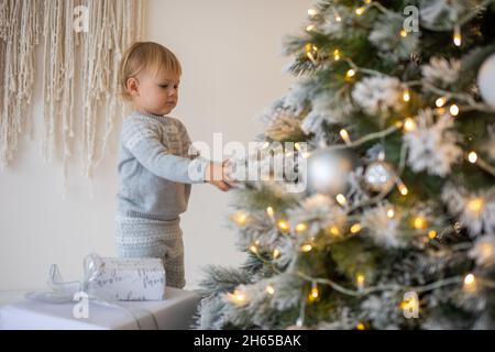 Adorable baby girl sitting near Christmas tree with festive lights and xmas gifts. Chrismas and New Year. Stock Photo