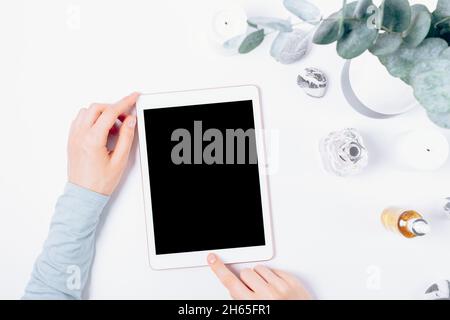 Female hands using tablet computer next to feminine accessories and eucalyptus branches on white table, top view. Stock Photo