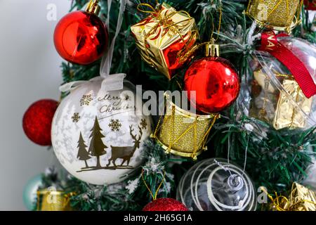 Close up of white Christmas bauble with the words Merry Christmas, hung on a Christmas tree along with other decorations, red baubles and gold gifts Stock Photo