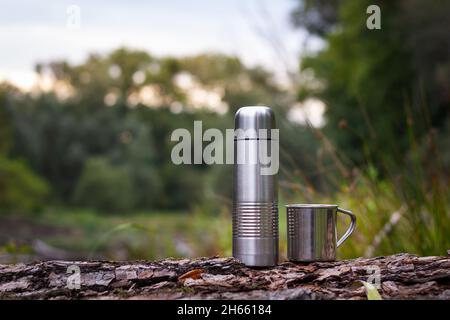 Thermos and stainless steel mug outdoors. Camping equipment on tree trunk in nature. Metal bottle and cup for hot drink Stock Photo