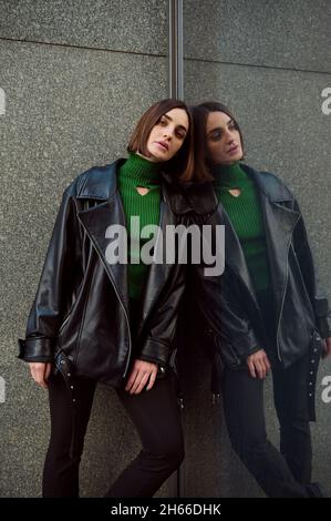 Attractive fashionable brunette woman in eco-leather clothes posing for the camera, leaning against the marble wall of a modern building reflected onA Stock Photo