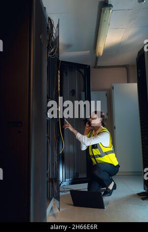 Caucasian female engineer with laptop talking on smartphone while inspecting in computer server room Stock Photo