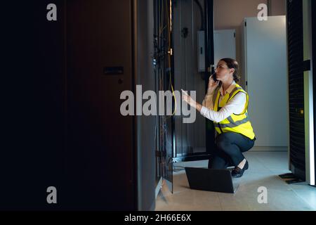 Caucasian female engineer with laptop talking on smartphone while inspecting in computer server room Stock Photo
