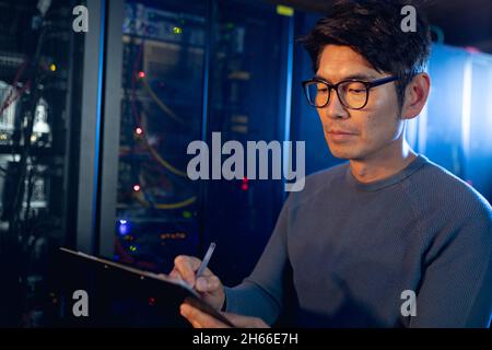 Asian male engineer writing on clipboard in computer server room Stock Photo