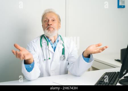 Portrait of mature adult male doctor in white uniform coat with stethoscope speaking, looking at camera, gesturing spreads arms out to sides. Stock Photo