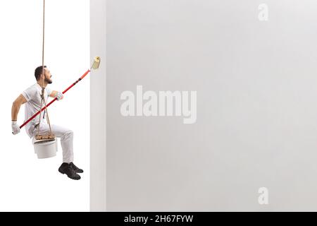 Side shot of a painter sitting on a swing and painting a wall with a paint roller isolated on white background Stock Photo