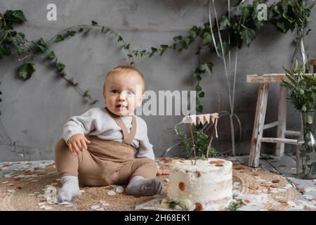 holiday baby's first birthday, boy with a delicious cake sitting on the floor, birthday in rustic style, natural photo zone, happy one year old. Stock Photo