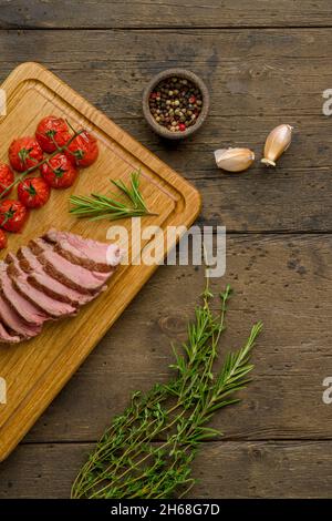 Grilled juicy and tender striploin slices with cherry tomatoes, thyme, rosemary, garlic and pepper on wooden board. Gourmet roasted meat. Culinary gas Stock Photo