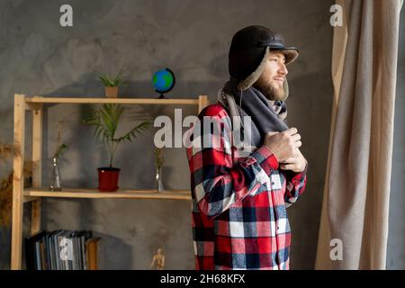 Heating is turned off for non-payment, a bearded Caucasian man stands in an apartment wearing a winter hat and a scarf. Cold winter. Stock Photo