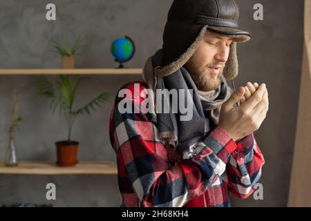 In winter, in a cold apartment without heating. A young Caucasian man wearing a hat and a scarf stands near the window and freezes. Close up portrait. Stock Photo