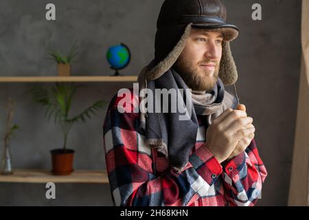 In winter, in a cold apartment without heating. A young Caucasian man wearing a hat and a scarf stands near the window and freezes. Close up portrait. Stock Photo