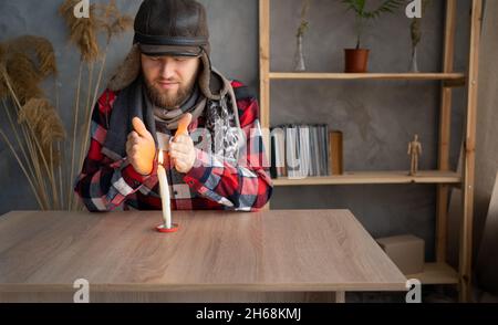 A Caucasian bearded man dressed in a warm winter hat and a scarf sits at home at a table and warms his hands from a burning candle. Stock Photo