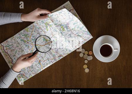 Map and magnifier in female hands on a wooden background, top view. Stock Photo