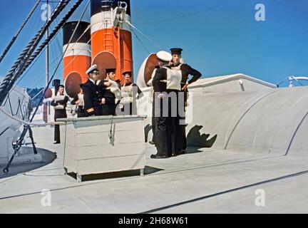 Uniformed officers and merchant sailors testing lifejackets or personal flotation devices onboard a freight ship of the Canada Steamship Lines (CSL) in 1955. The sailor on the left wears one marked ‘Front Childs’. The canvas pouch contained flexible waterproof cells filled with kapok. This device (PFD, life jacket, life preserver, life belt, Mae West, life vest, life saver, cork jacket, buoyancy aid or flotation suit) – it is worn to prevent drowning. Canada Steamship Lines is a shipping company with headquarters in Montreal, Quebec, Canada – a vintage 1950s photograph. Stock Photo