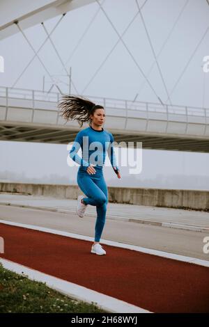 Pretty young woman in blue track suit running by the river at autumn morning Stock Photo