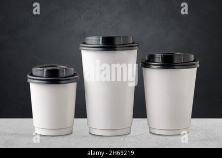 Three different sizes of white takeaway paper cups with black plastic lids isolated on gray background. Stock Photo