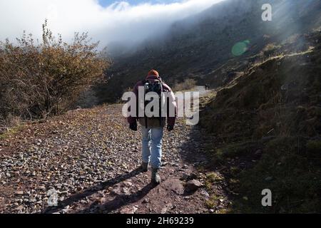 A mountaineer, senior adult man, in his 70s, wearing a down coat and backpack, walks down a dirt track towards the cloudy mountains, Pyrenees, Spain Stock Photo