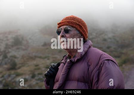 A mountaineer, senior adult man, in his 70s, wearing a down coat, gloves and sunglasses, enjoys the scenery during a mountain hike, Pyrenees, Spain Stock Photo
