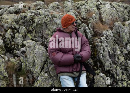 A mountaineer, senior adult man, in his 70s, in a feather coat, enjoys the scenery while resting leaning on some rocks full of lichen, Pyrenees, Spain Stock Photo