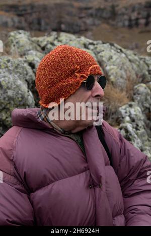 A mountaineer, senior adult man, in his 70s, in a feather coat, enjoys the scenery while resting leaning on some rocks full of lichen, Pyrenees, Spain Stock Photo
