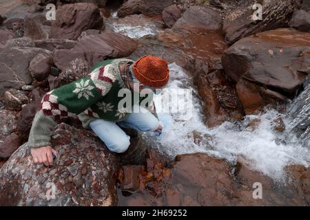 A mountaineer, senior adult man, in his 70s, wearing a wool sweater, stands next to a stream of water, collecting water with some water jugs, Pyrenees Stock Photo