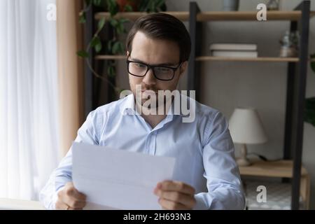 Concentrated young male manager in glasses reading paper document. Stock Photo