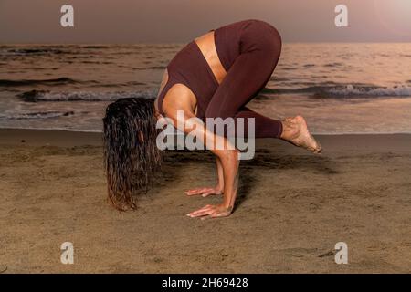 Woman practicing yoga on the beach at sunset. Woman in the Kakasana position, crow position for concentration in the present. Stock Photo