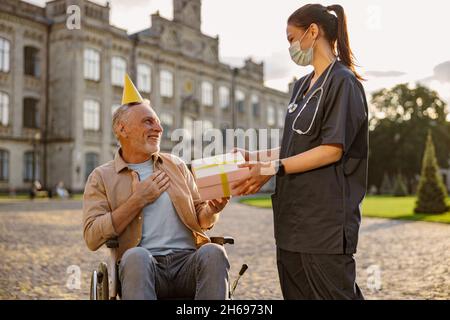 Pleased mature man, recovering patient in wheelchair wearing birthday hat receiving gift and congratulations from young nurse Stock Photo