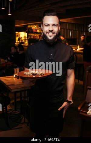Cheerful bearded waiter with a slice of cake in a Georgian restaurant. Beautiful waiter in black clothes with a beard and a serving of cake Stock Photo