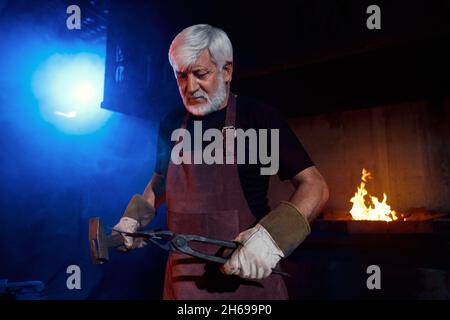 Aged caucasian blacksmith in protective apron and gloves using hammer and forceps while working at forge. Bearded man processing molten metal indoors. Manual work.  Stock Photo