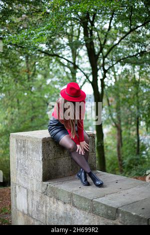 Looking down white woman with long dark hair in bright red sitting on stone wall in park Stock Photo